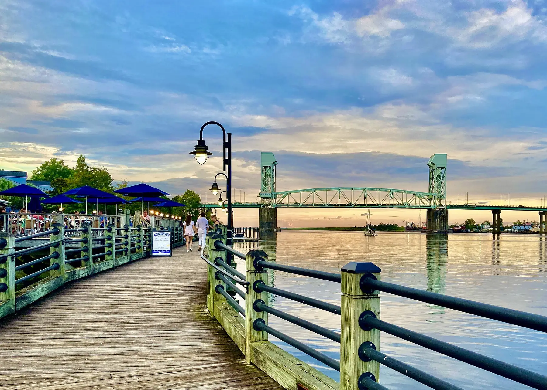 The Riverwalk along the Cape Fear River in downtown Wilmington at sunset, with the Cape Fear Memorial Bridge beyond.