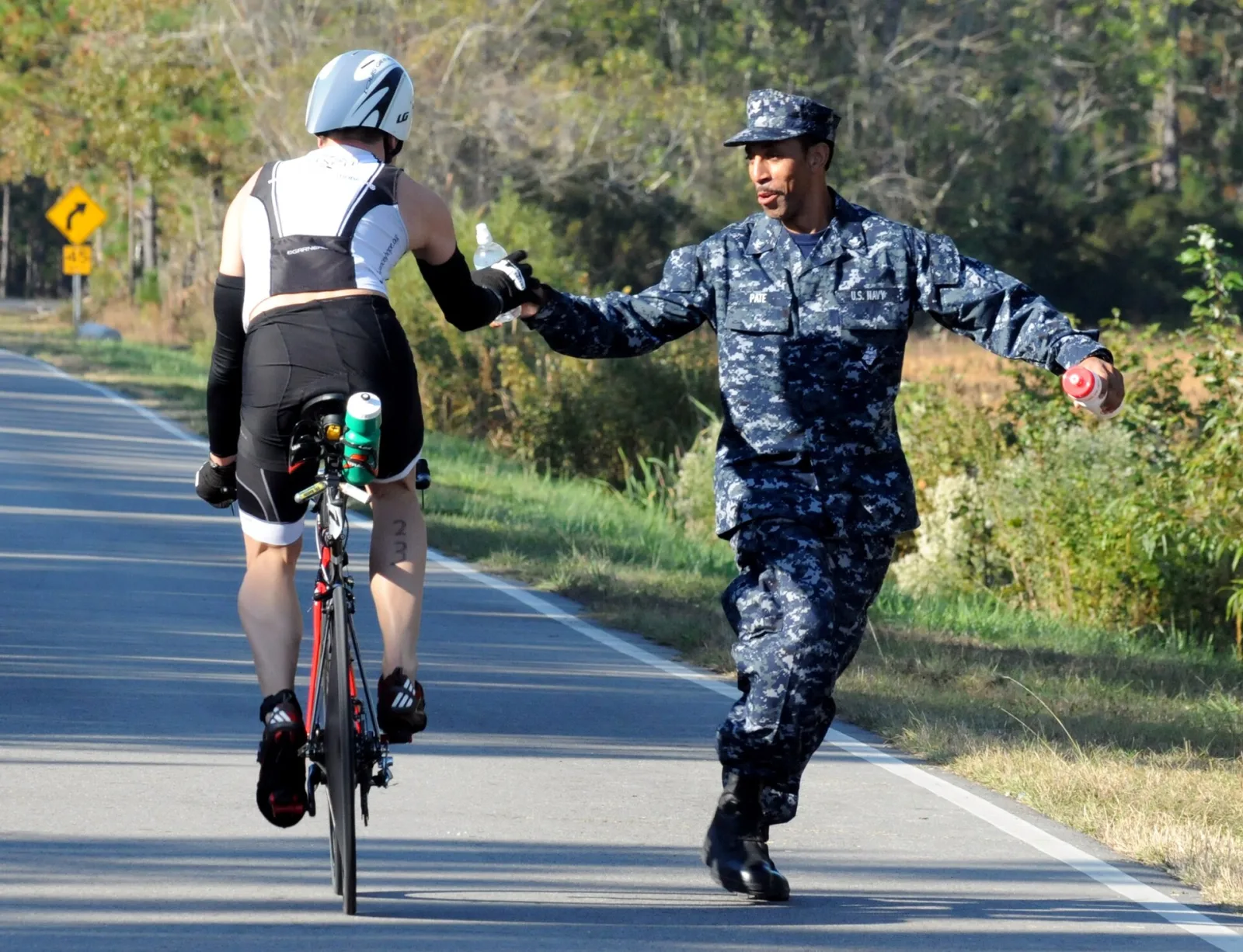 A U.S. Navy sailor in uniform runs alongside a cyclist on a country road and hands over a water bottle during a triathlon near Wilmington.